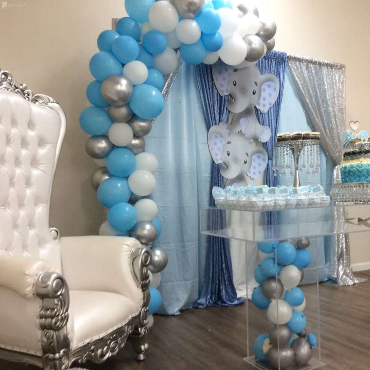 The scene features a decorated party setup with a balloon arch, a white throne chair, and a dessert table filled with cupcakes and treats in blue and silver tones.