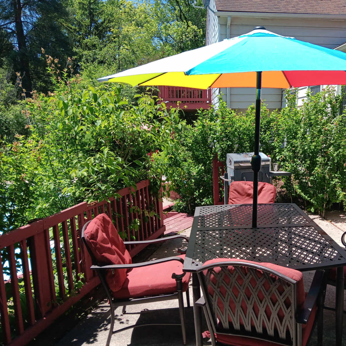 A table with four chairs is covered by a colorful umbrella on a patio surrounded by greenery.