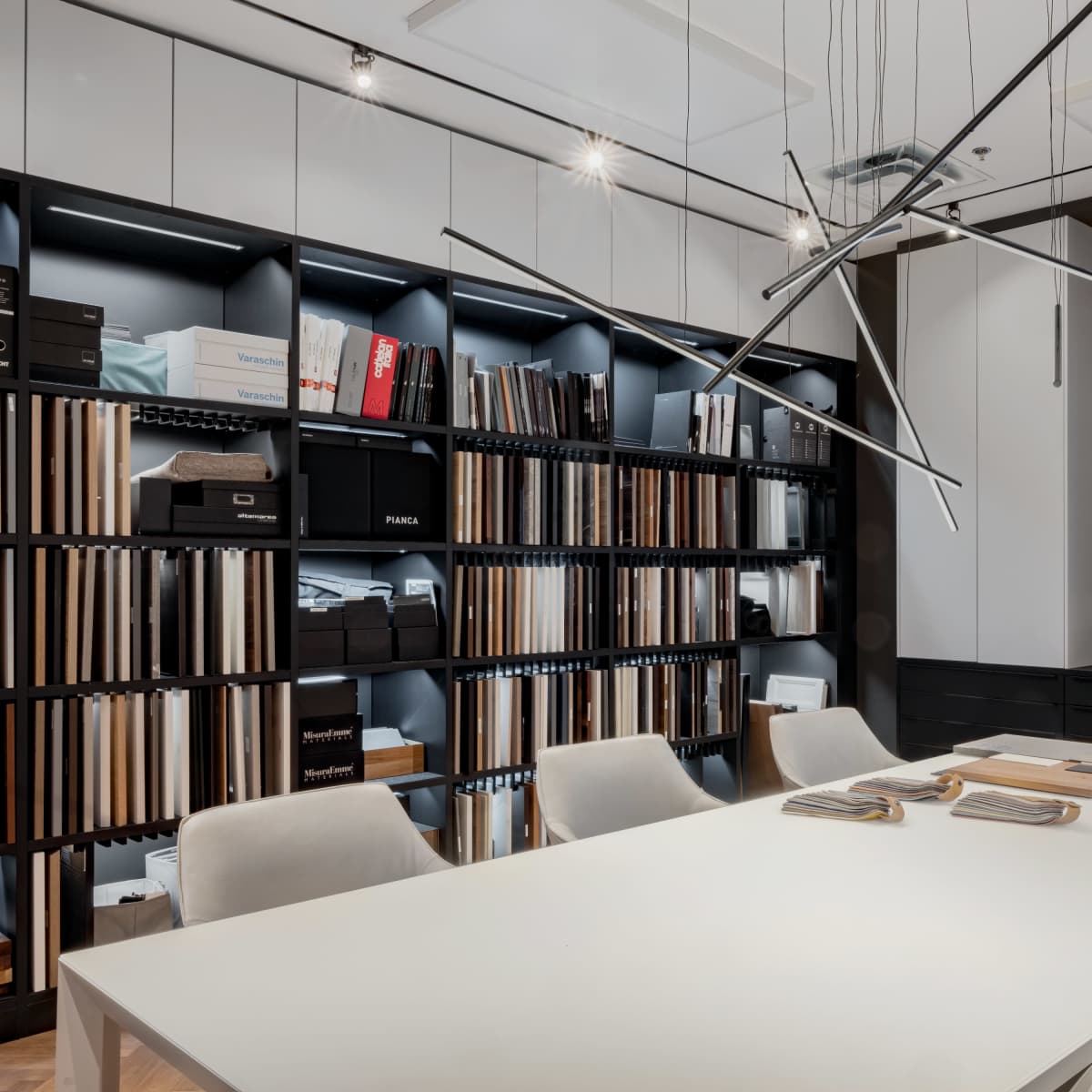 A modern office space features a large white table surrounded by chairs, flanked by a wall of organized file storage and a television screen displaying a room design.