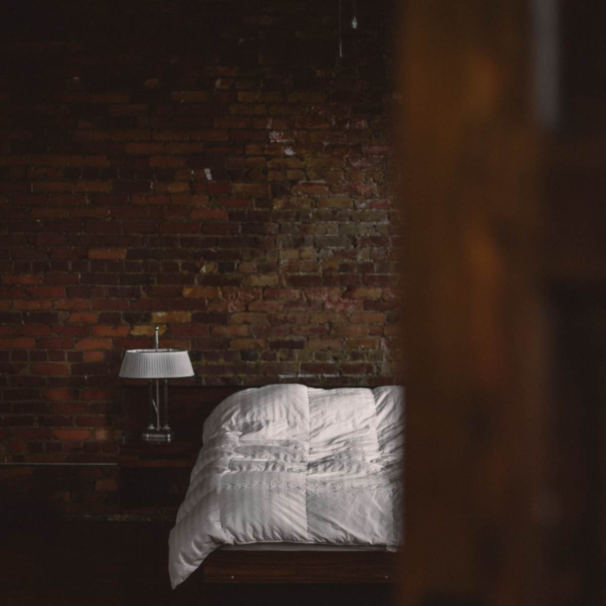 A neatly made bed with a white comforter is positioned against a textured brick wall in a dimly lit room.