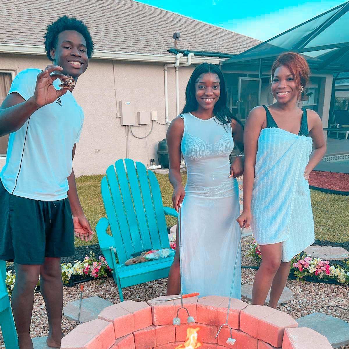 Three people stand around a fire pit, each holding a skewer, in a backyard setting with colorful chairs and flowers in the background.
