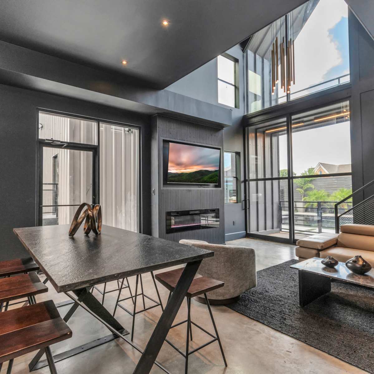 A modern living space with a high ceiling, large windows, a dining table, and a sectional sofa, overlooking an outdoor view.