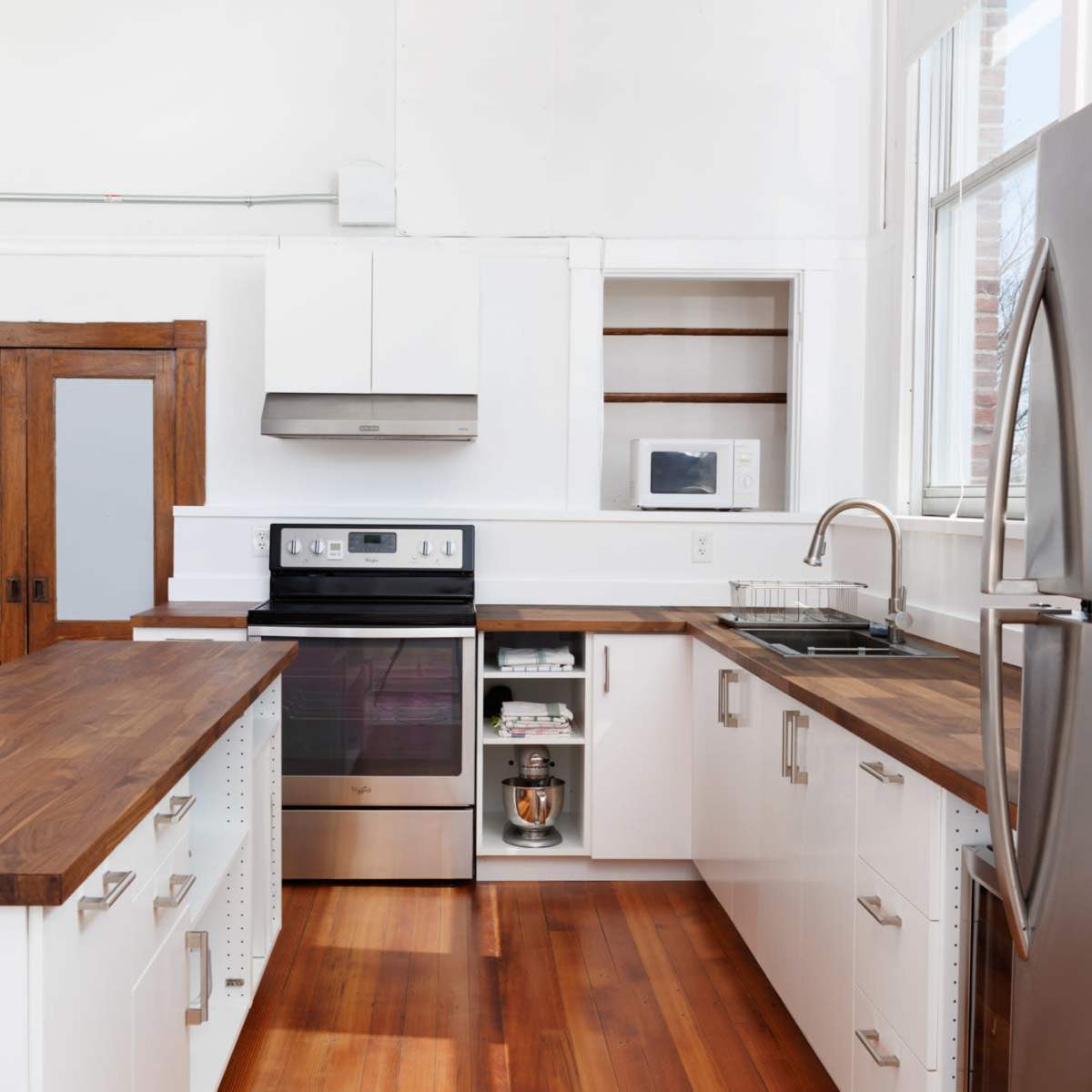 A modern kitchen featuring white cabinets, a stainless steel refrigerator, an oven, and wooden countertops, with large windows allowing natural light.