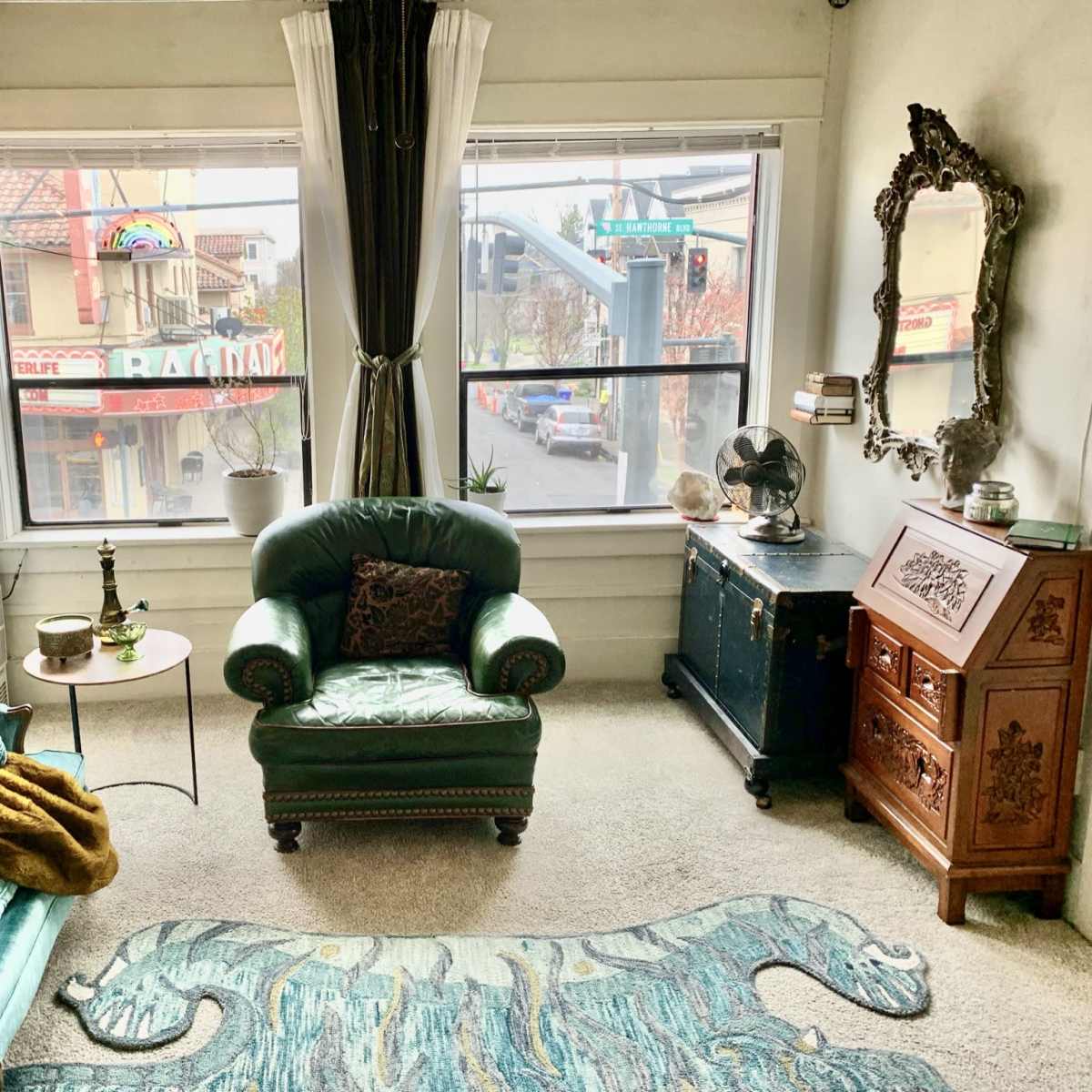 The room features a green velvet sofa and armchair, a decorative rug, and large windows with a view of a street lined with shops.