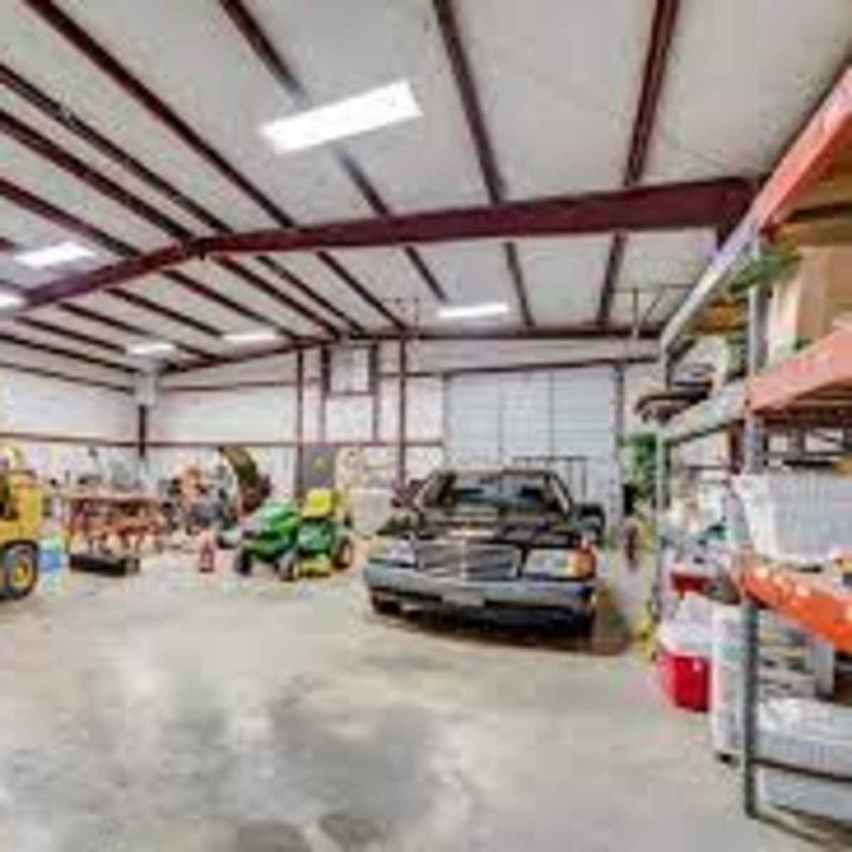 A spacious industrial garage with various equipment, including a forklift, lawnmower, and a parked car, along with shelves filled with tools and supplies.