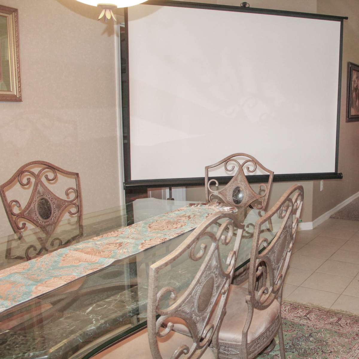 A dining area with a glass table and four chairs, a pull-down projector screen, and a small couch in a room with beige walls and carpet.