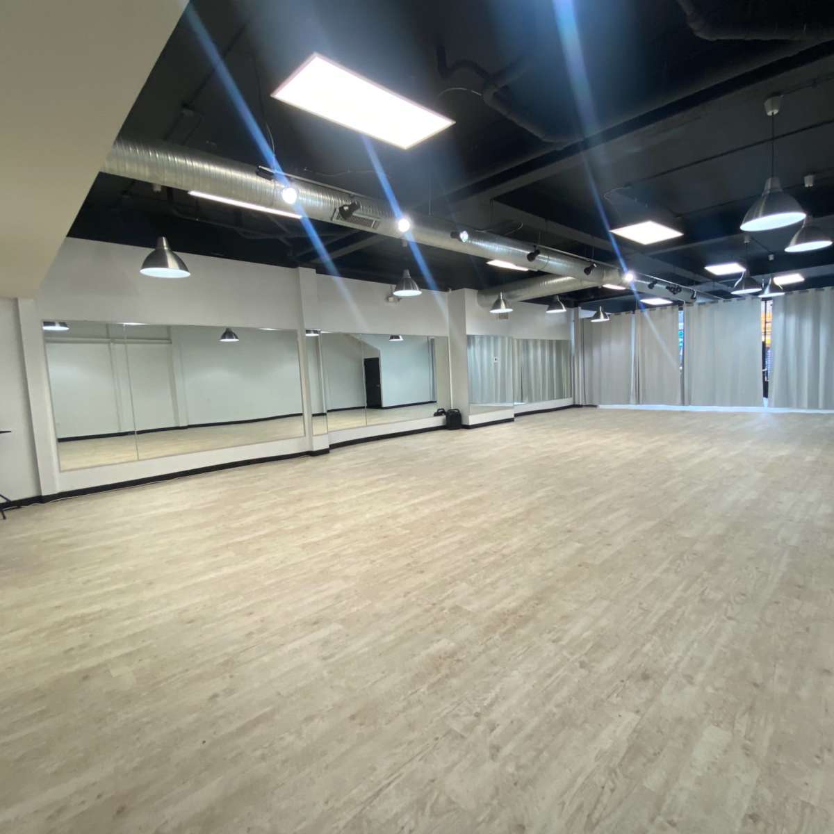 A spacious, empty dance studio with mirrored walls, wooden flooring, and industrial-style lighting fixtures.