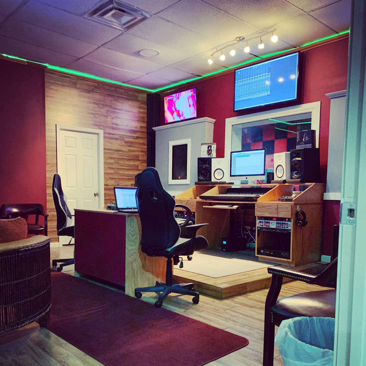A recording studio with a control desk, monitors, and audio equipment, featuring wooden accents and a red wall.