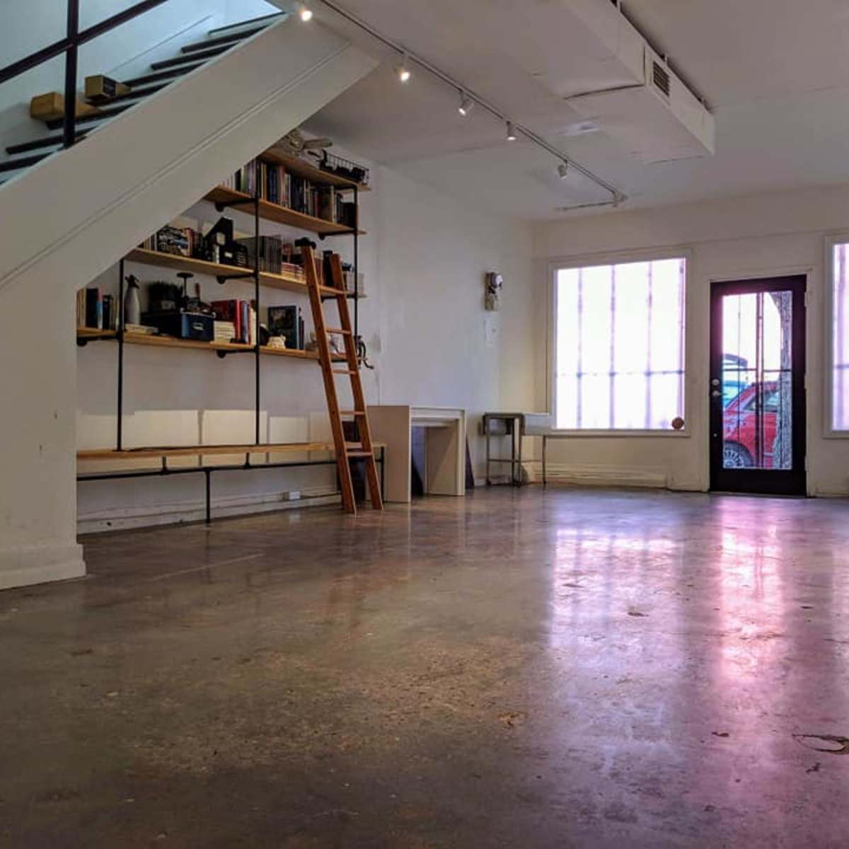 A spacious, empty room with polished concrete flooring, shelves filled with various items against one wall, and large windows allowing natural light to enter.