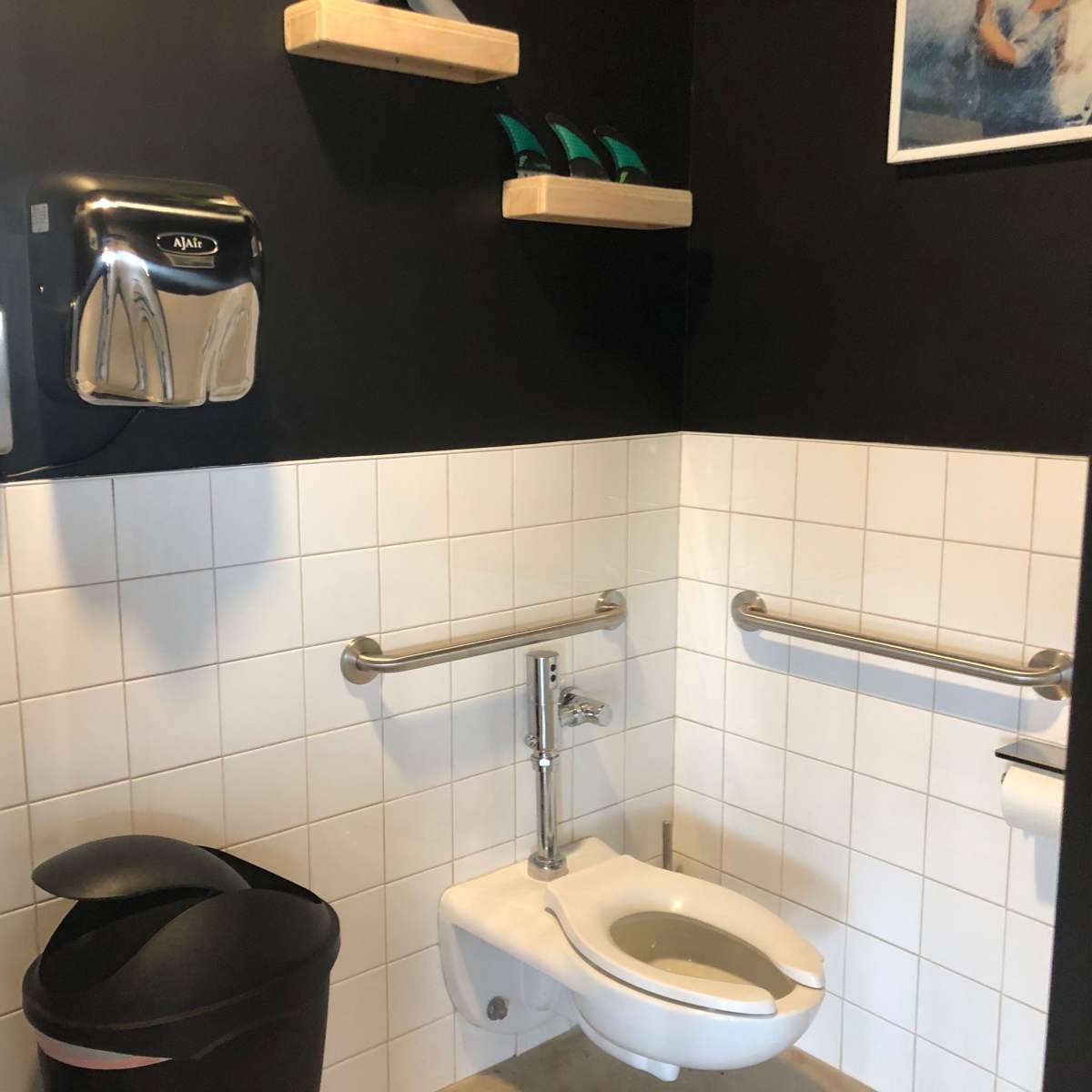 A small bathroom featuring a toilet, a sink, a paper towel dispenser, and grab bars along the walls, with a black accent wall and white tiled sections.