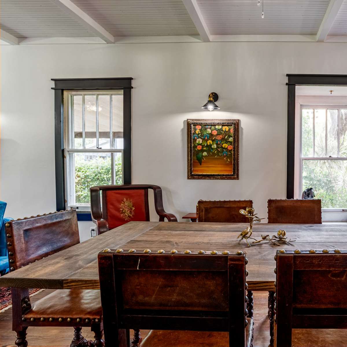 A wooden dining table with six chairs is set in a well-lit room featuring a window and a piece of framed artwork on the wall.