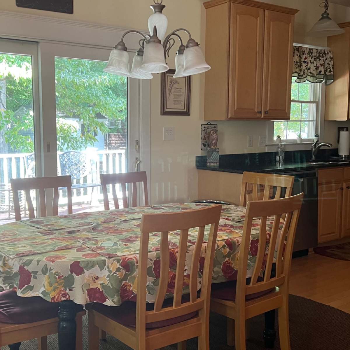 A dining area features a wooden table covered with a floral tablecloth surrounded by six chairs, with sliding glass doors leading to a deck in the background.
