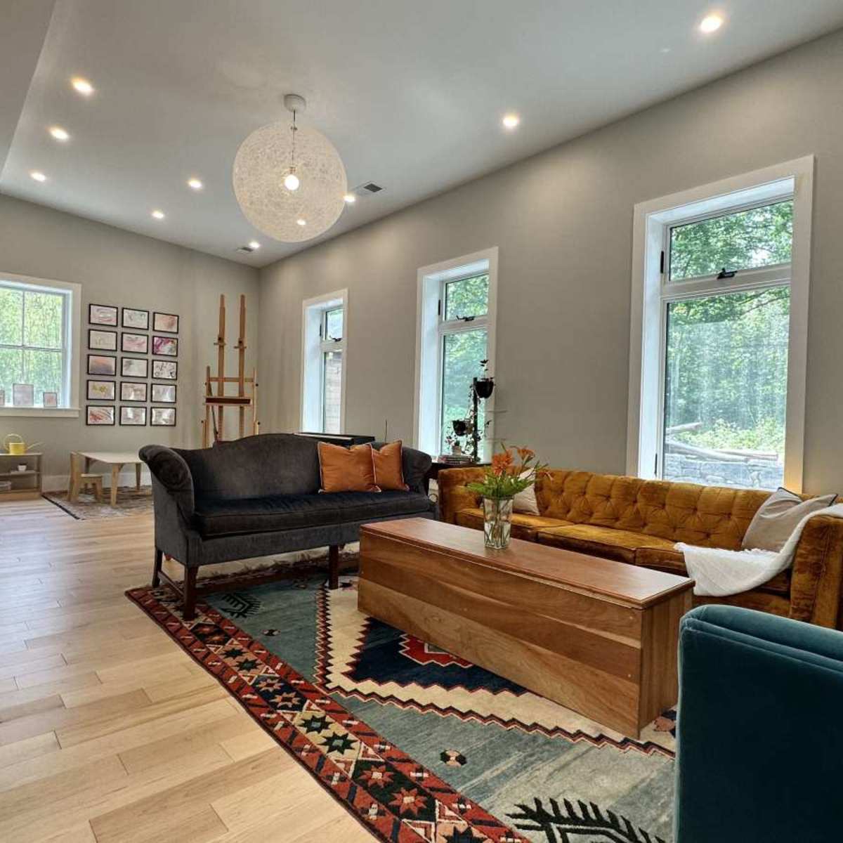 A spacious living room with a wooden floor, featuring a mix of furniture including a yellow couch, a dark sofa, and a wooden coffee table, along with large windows that let in natural light.
