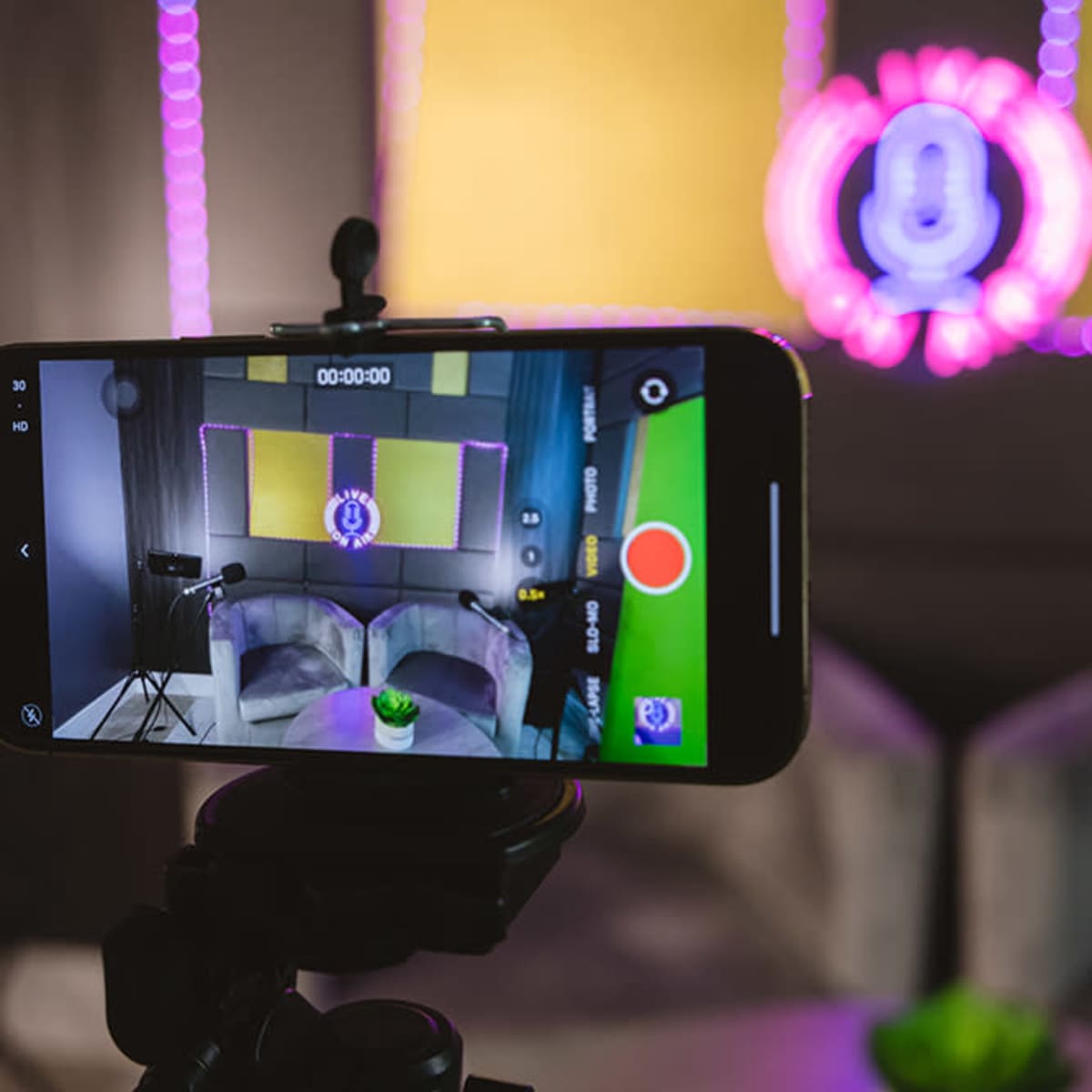 A smartphone on a tripod captures a recording setup featuring two chairs, a small plant, and a backdrop with purple lighting.