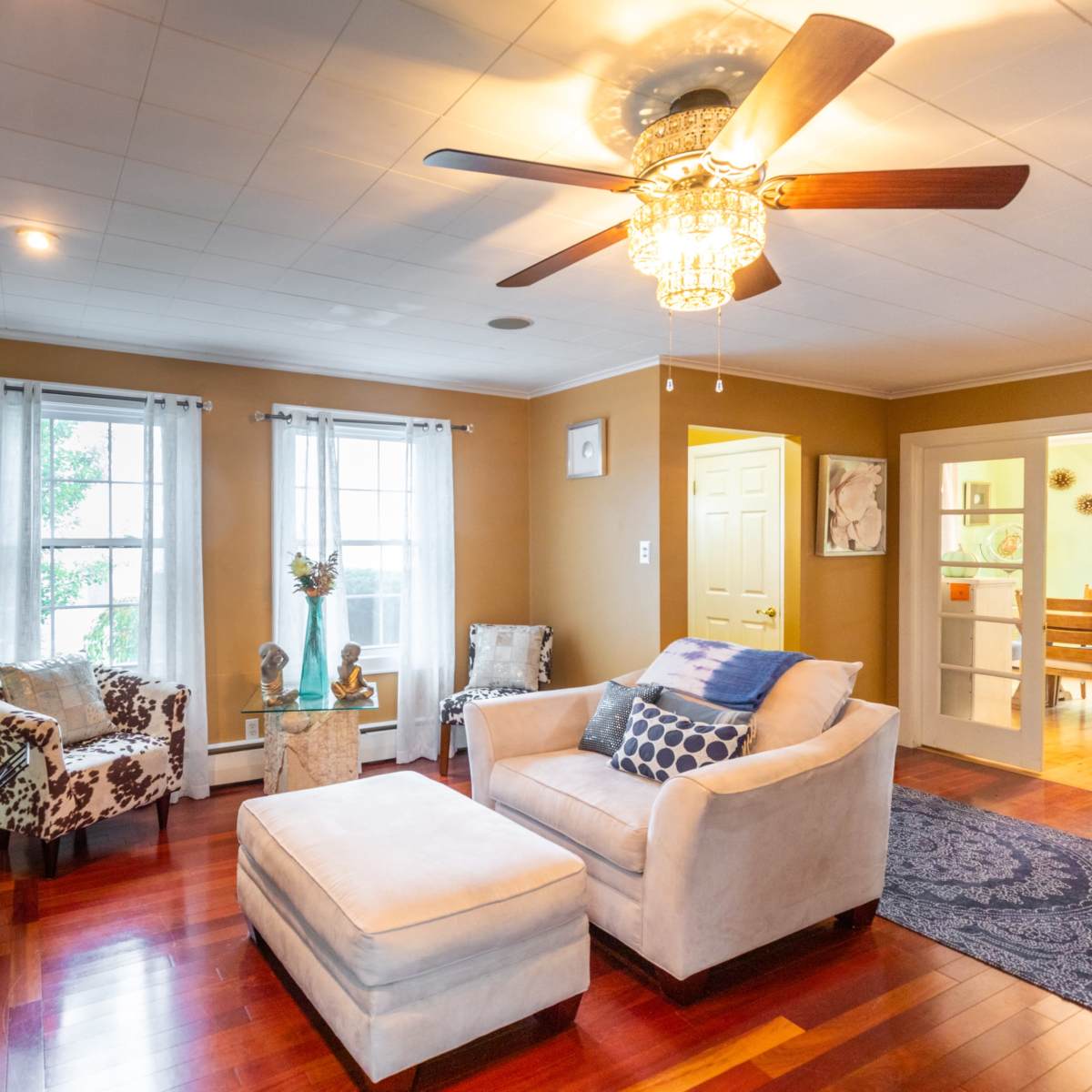 A cozy living room with a ceiling fan, two chairs, a sofa, and a hardwood floor, complemented by natural light from windows.