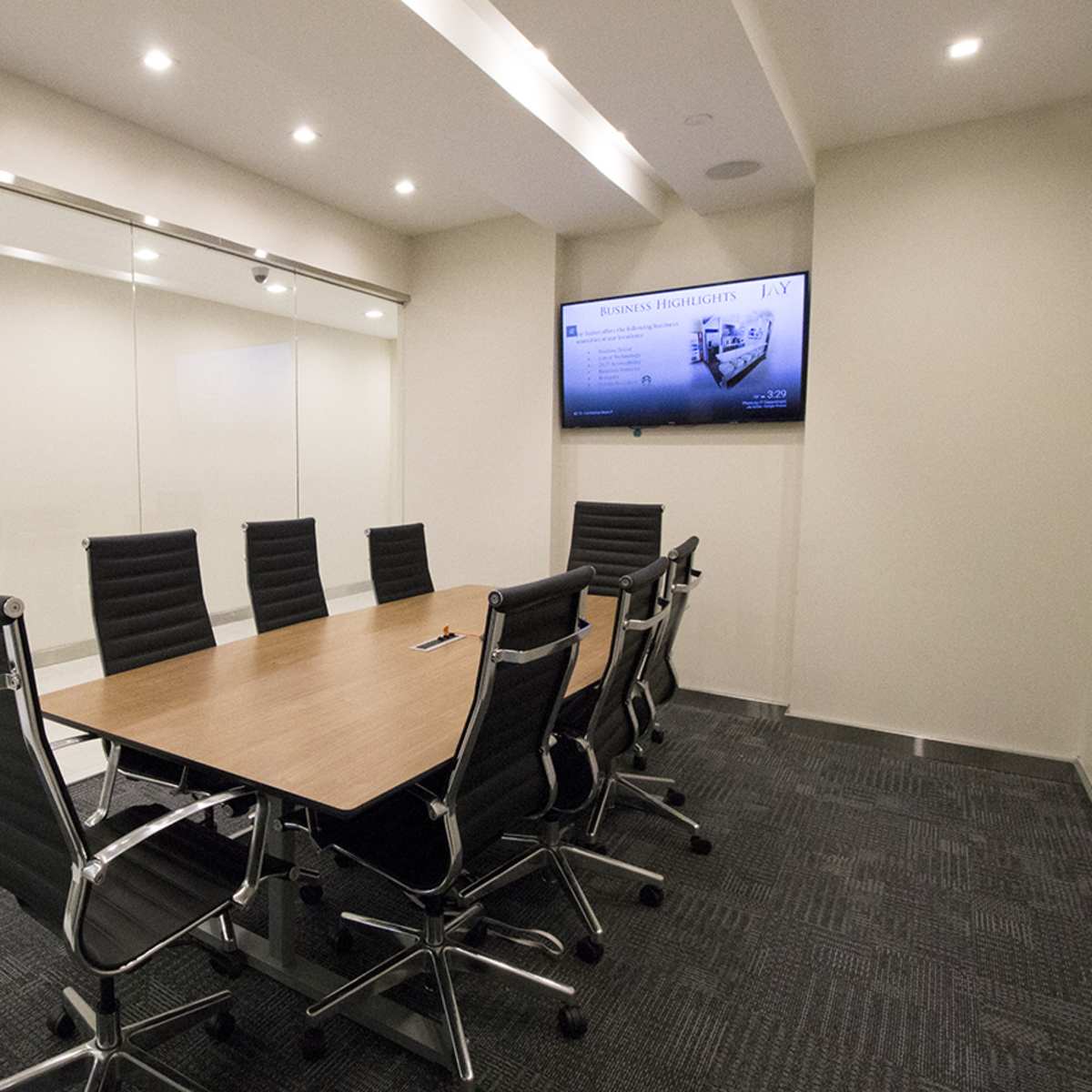 A conference room features a large wooden table surrounded by black leather office chairs, with a wall-mounted screen and glass walls.