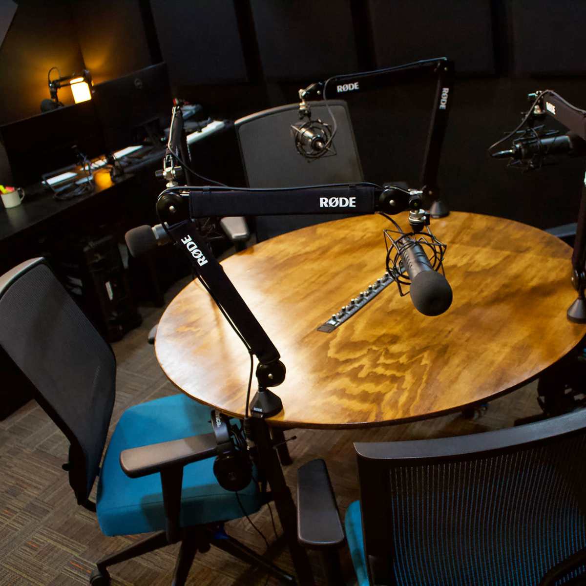 A round wooden table is set up in a recording studio with four adjustable chairs and mounted microphones.