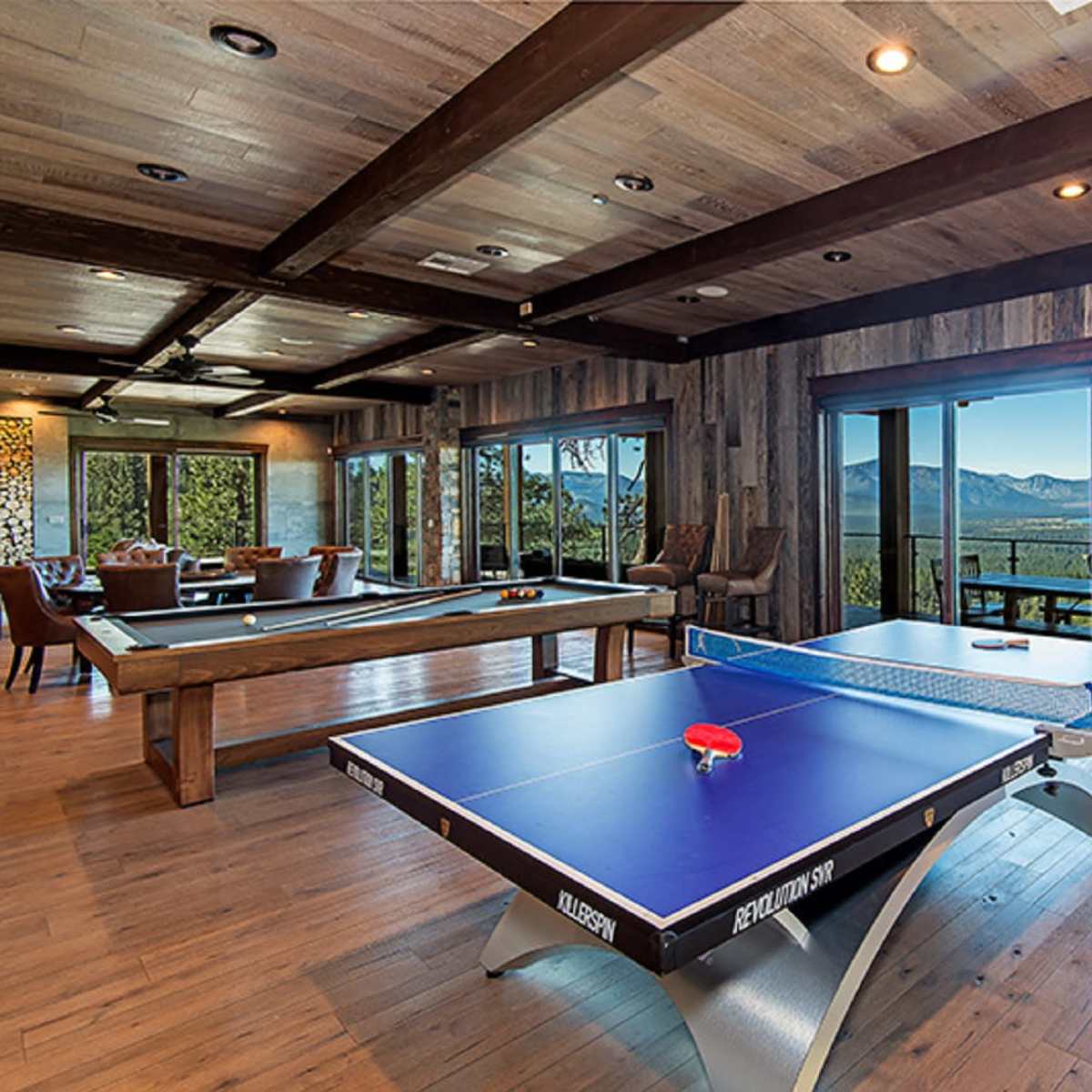 A game room featuring a ping pong table and a pool table, with large windows offering views of a lake and mountains in the background.