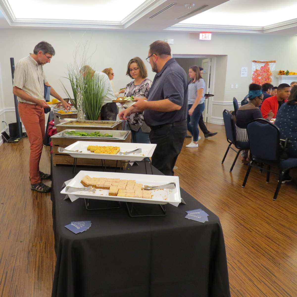 People are serving themselves food from a buffet table in a well-lit room with a gathering of seated attendees in the background.