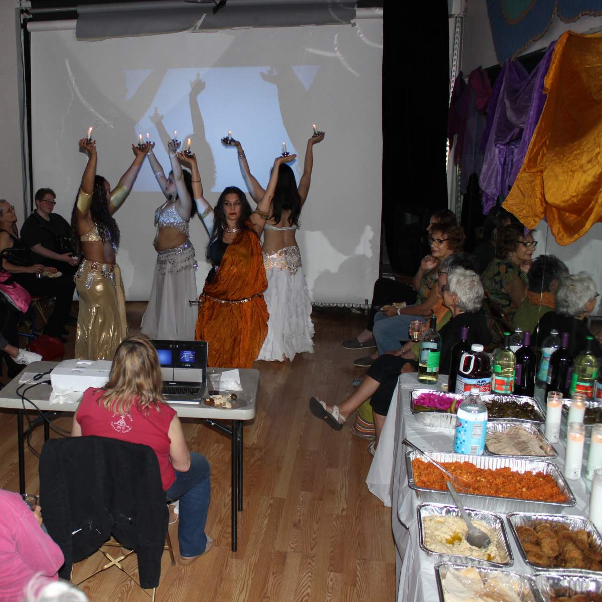 A group of dancers performs on stage while an audience watches, surrounded by tables with food and drinks.