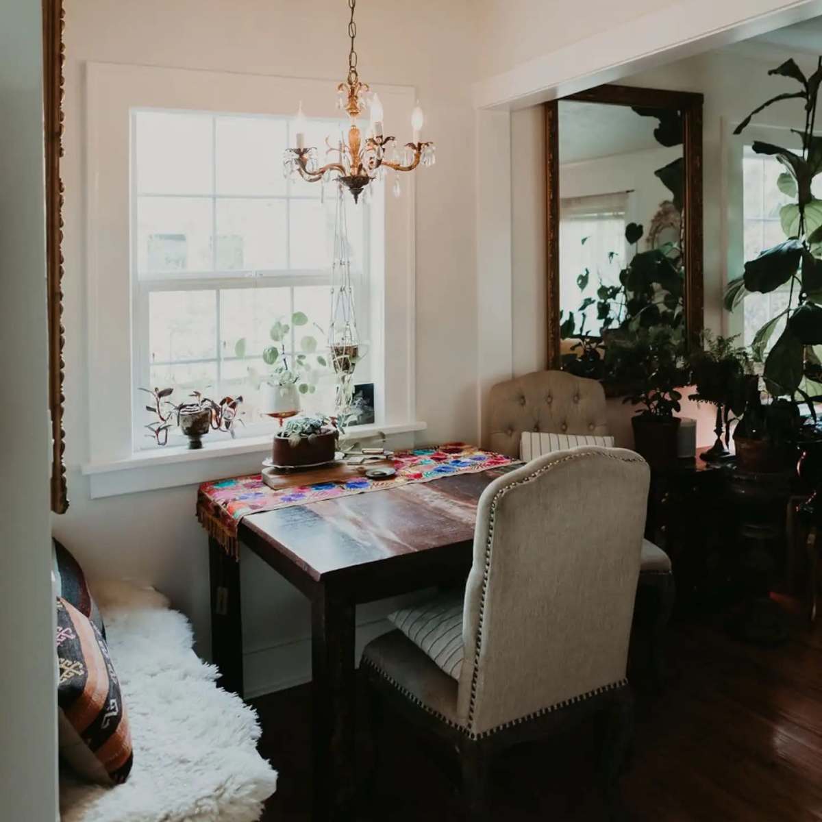 A small dining area features a wooden table with two chairs under a chandelier, next to large windows and surrounded by greenery.
