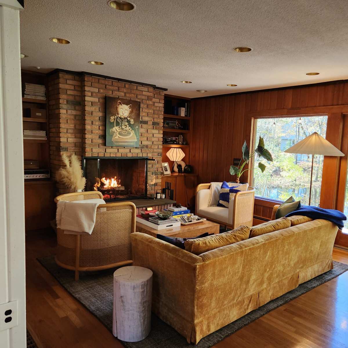 A cozy living room with a brick fireplace, a yellow sofa, wooden walls, a coffee table, and large windows overlooking trees.