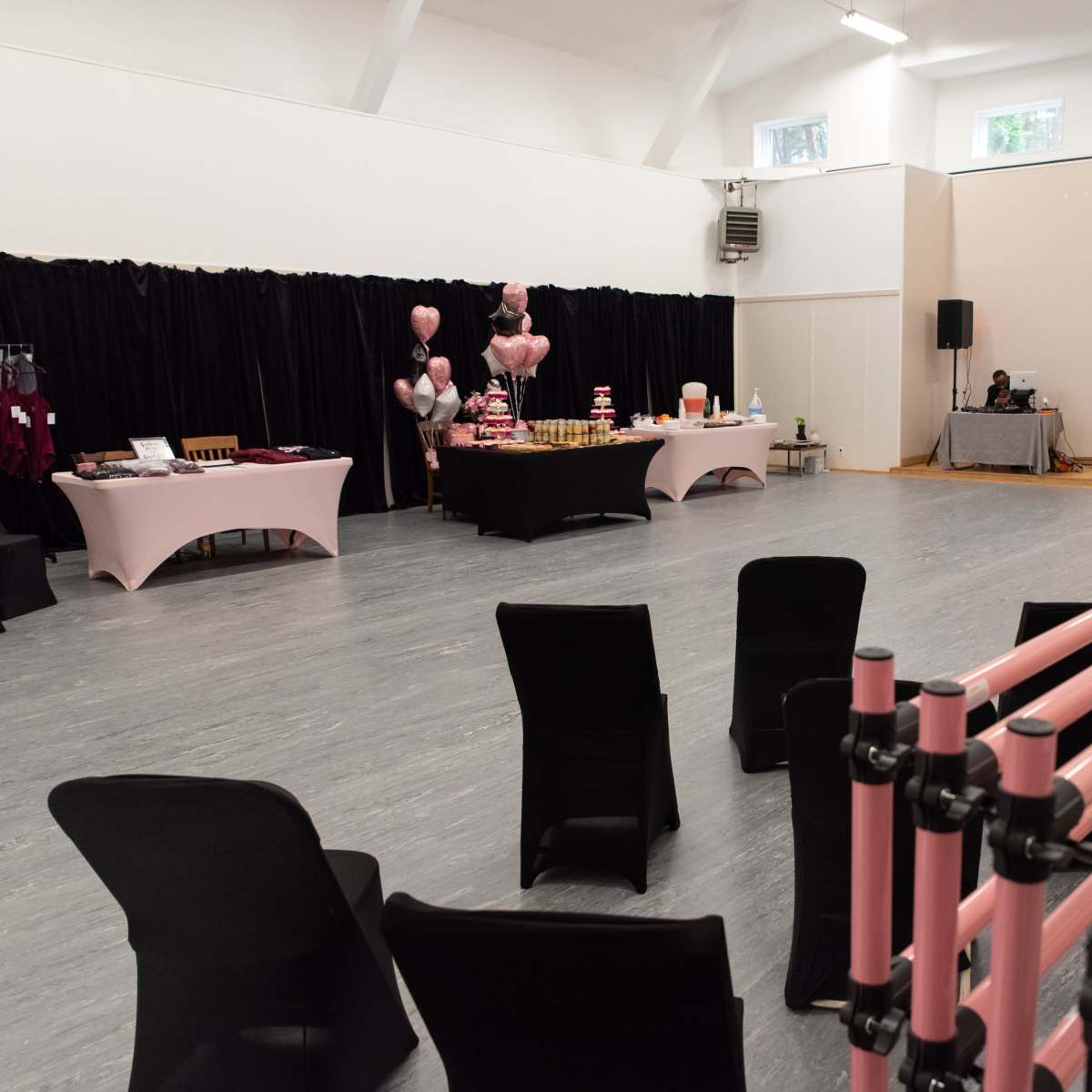 A spacious event room features rows of black chairs facing a table adorned with pink decorations, snacks, and a speaker setup in the background.