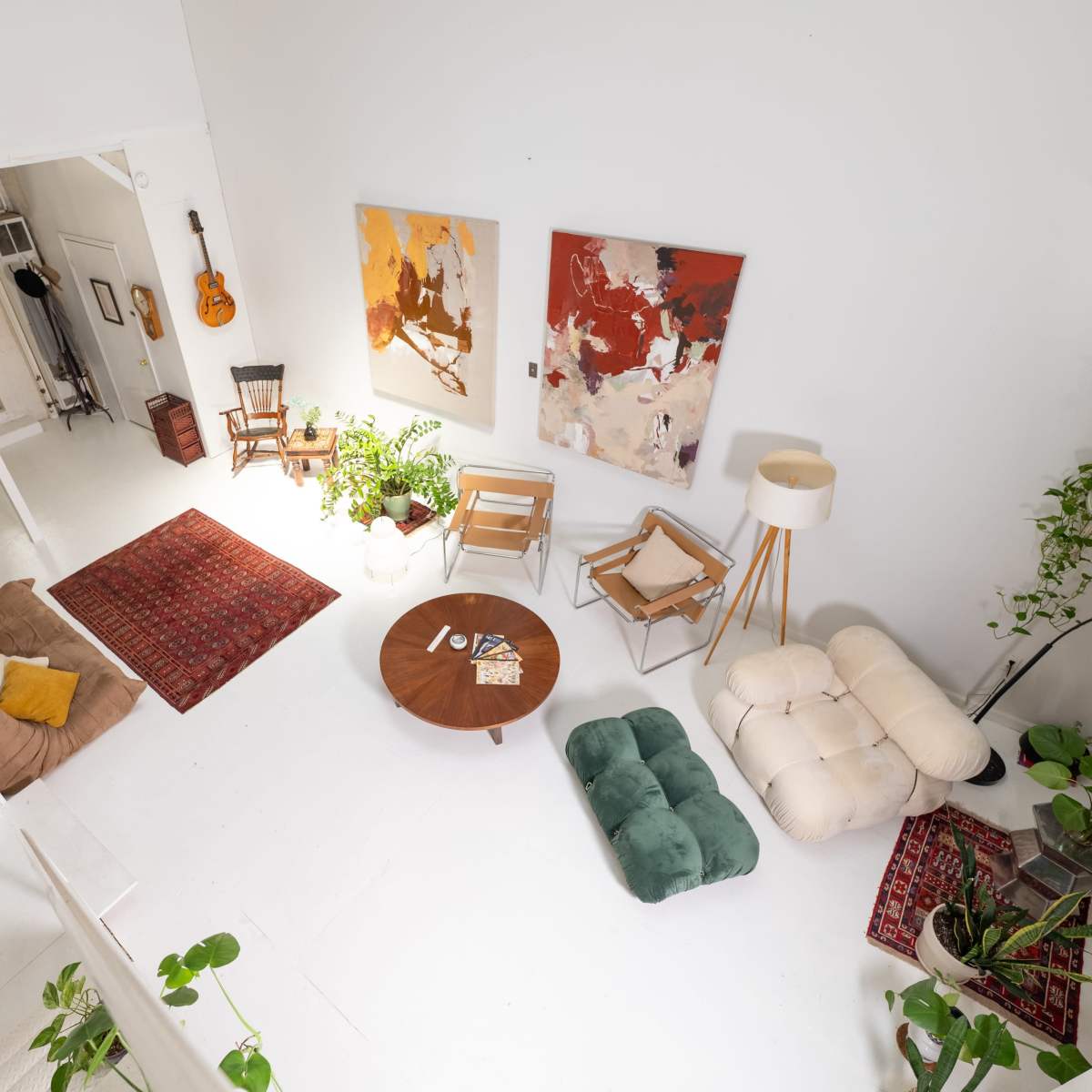 A bright, spacious living room features a mix of furniture, including a round table, sofas, and plants, with artwork on the walls and a staircase visible in the background.