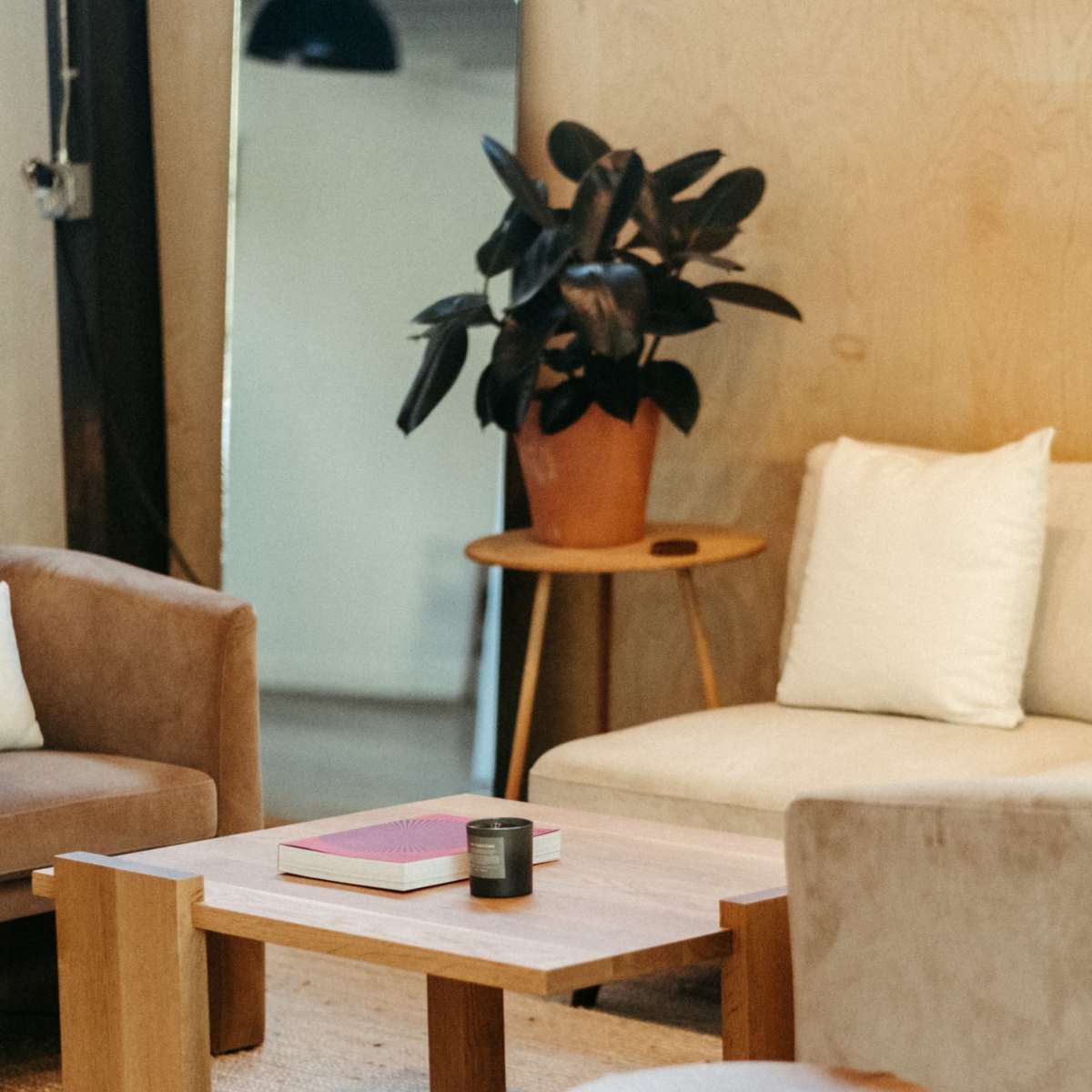 A minimalist living space features a wooden table with a book and a candle, surrounded by stylish sofas and a potted plant.