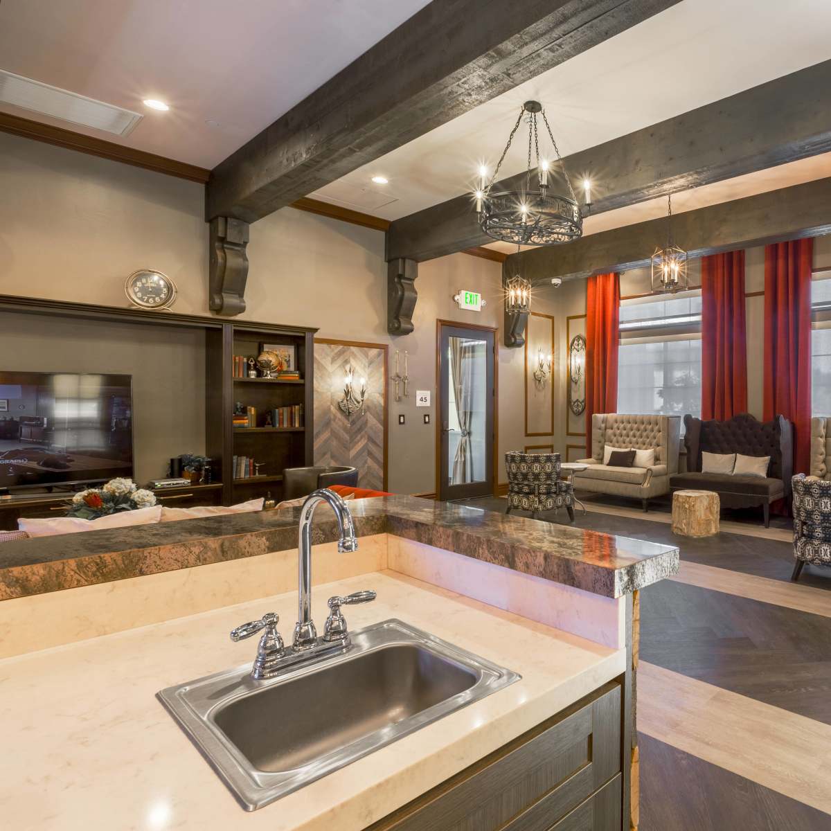 A modern living room with beige sofas, red curtains, and a kitchen area featuring a sink and granite countertop.