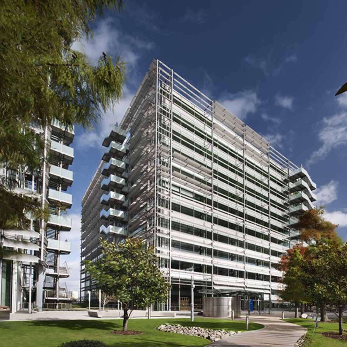 A modern glass building with multiple balconies, surrounded by green landscaping and a clear blue sky.