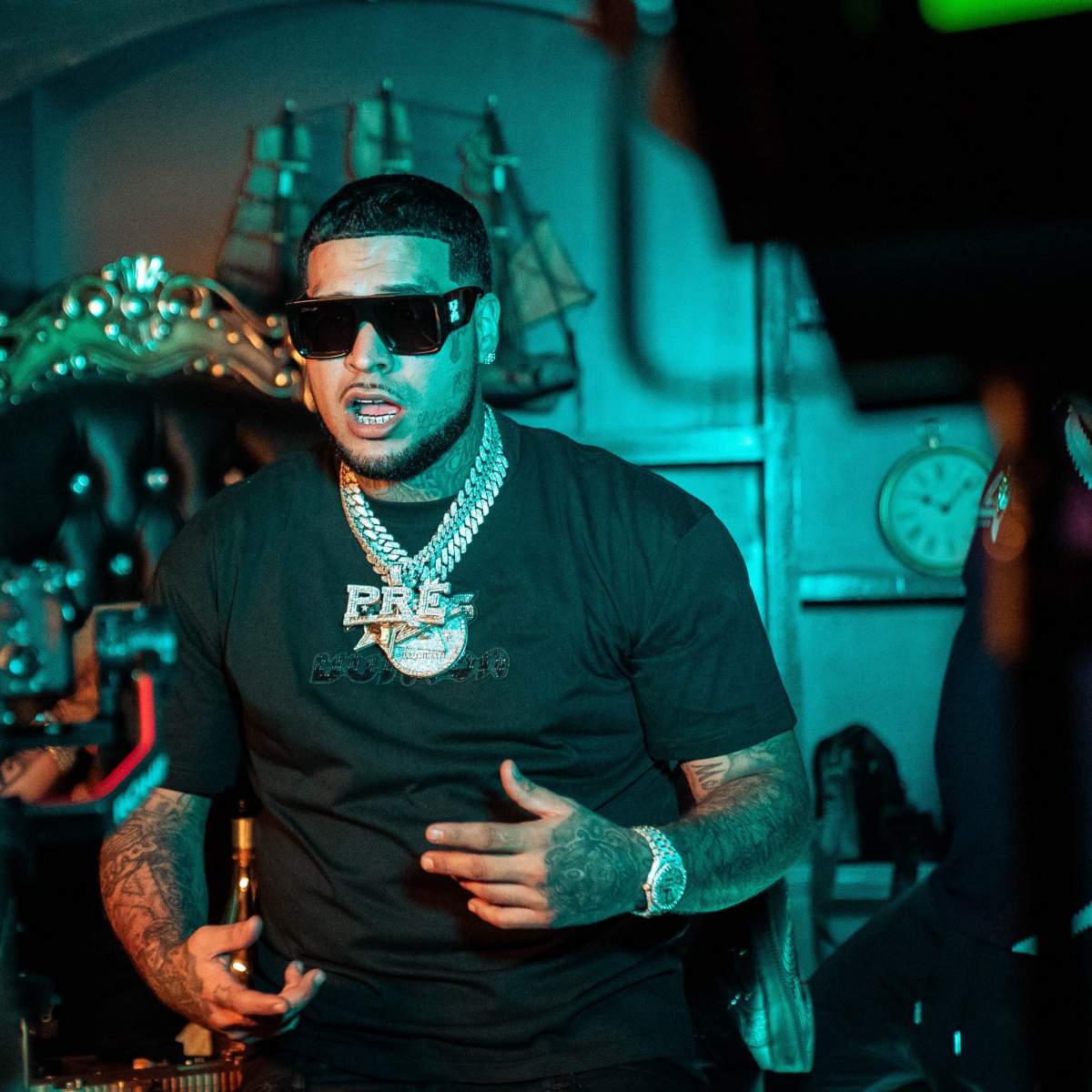 A man wearing sunglasses and jewelry is sitting in a dimly lit room while gesturing with his hands.