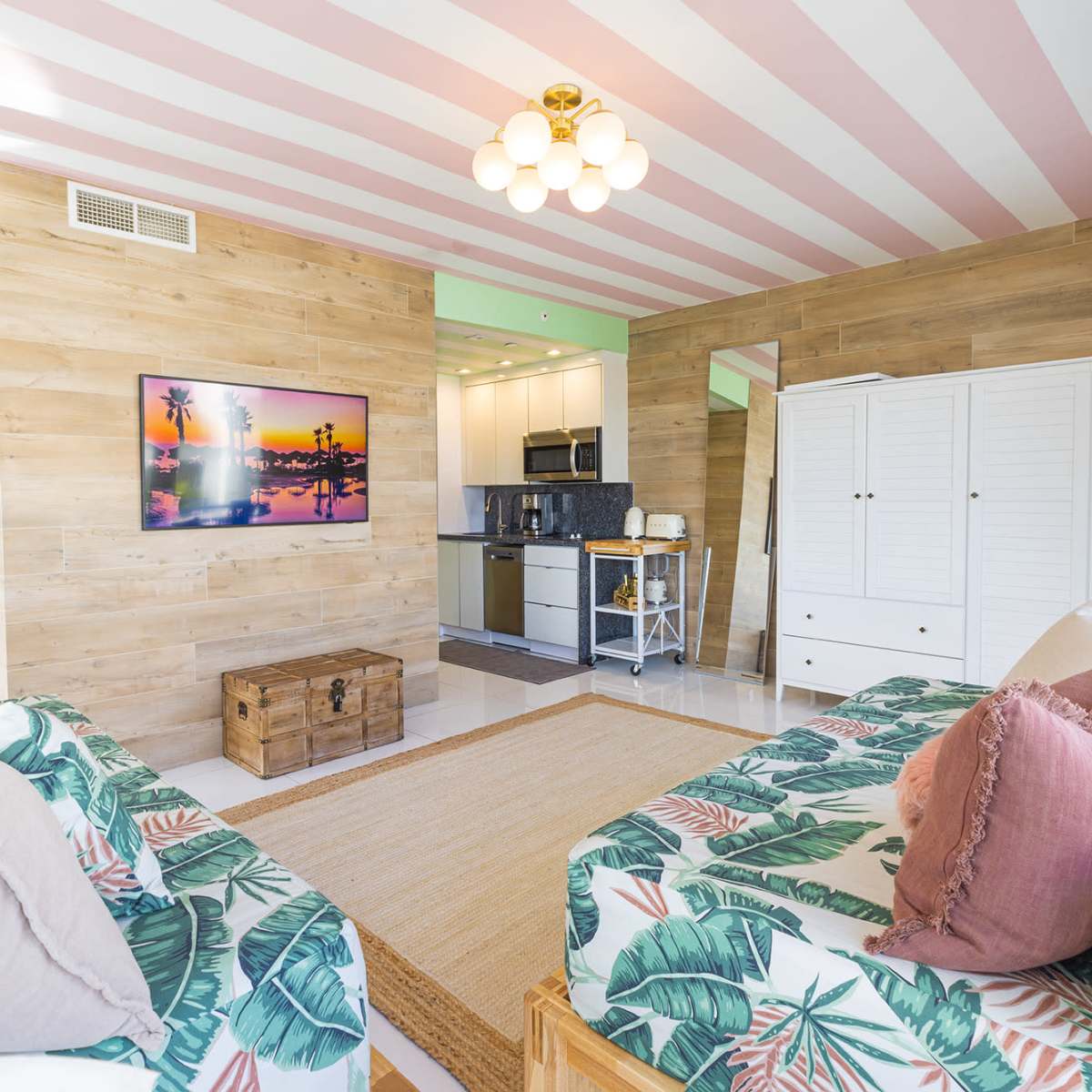 A brightly lit room features two patterned sofas, a wooden coffee table, a television mounted on a wood-paneled wall, and a kitchenette in the background.