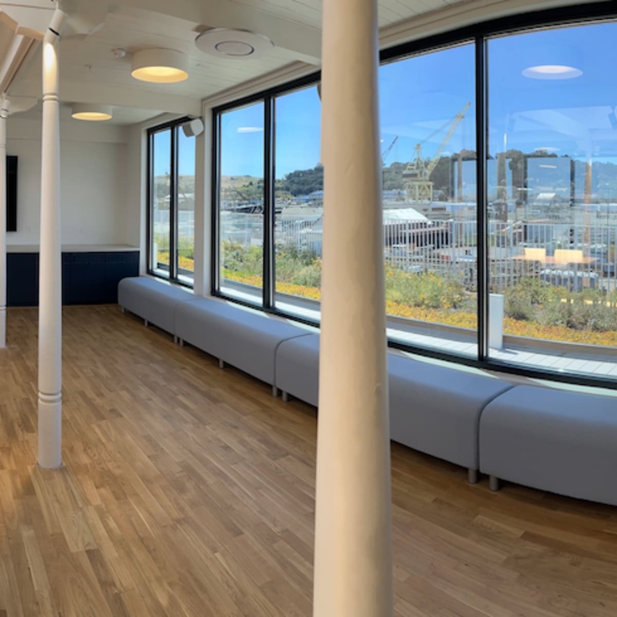A large, empty meeting room features a long wooden table surrounded by chairs, with large windows offering a view of an outdoor area and distant buildings.