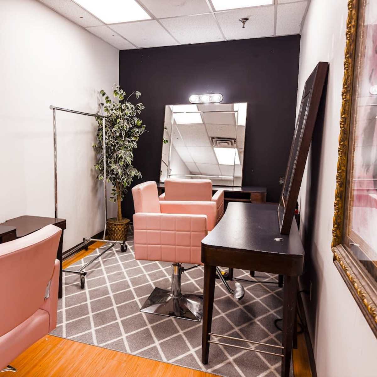 A cosmetology or salon room featuring two pink styling chairs, a mirror, and a clothing rack, all set in a space with a gray carpet and black walls.