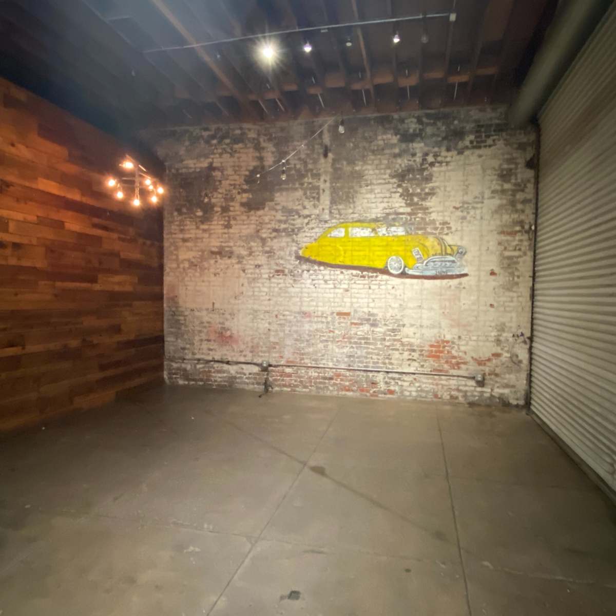 An empty industrial space with a brick wall, wooden paneling, and a mural of a yellow surfboard beside a large shutter door.