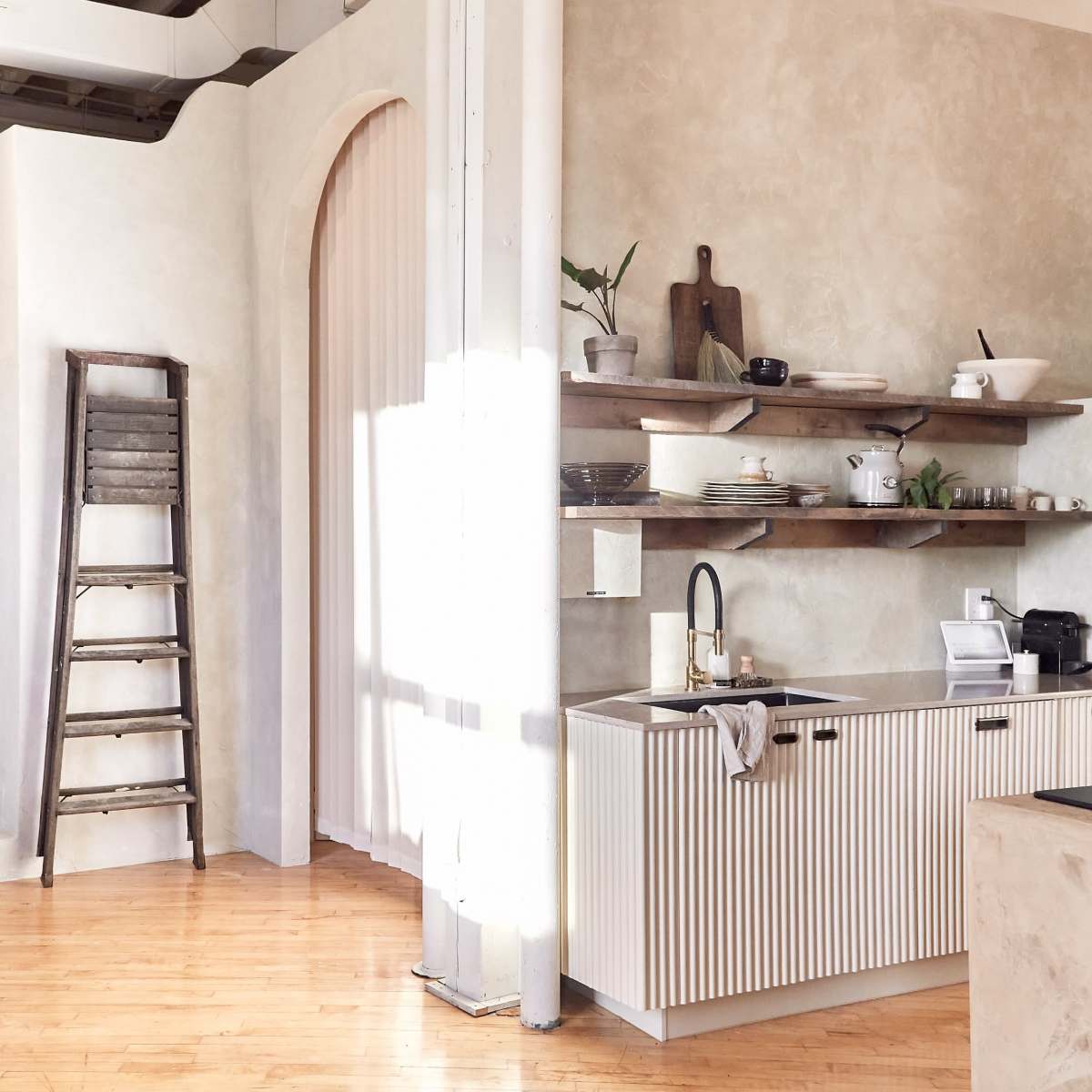 A minimalist interior space featuring a kitchen with a shelf and a ladder, alongside stairs leading to a different area with soft furniture.