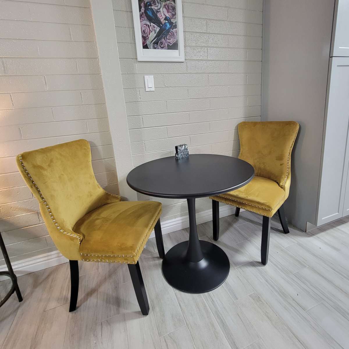 A round black table is positioned between two yellow upholstered chairs against a light gray brick wall.