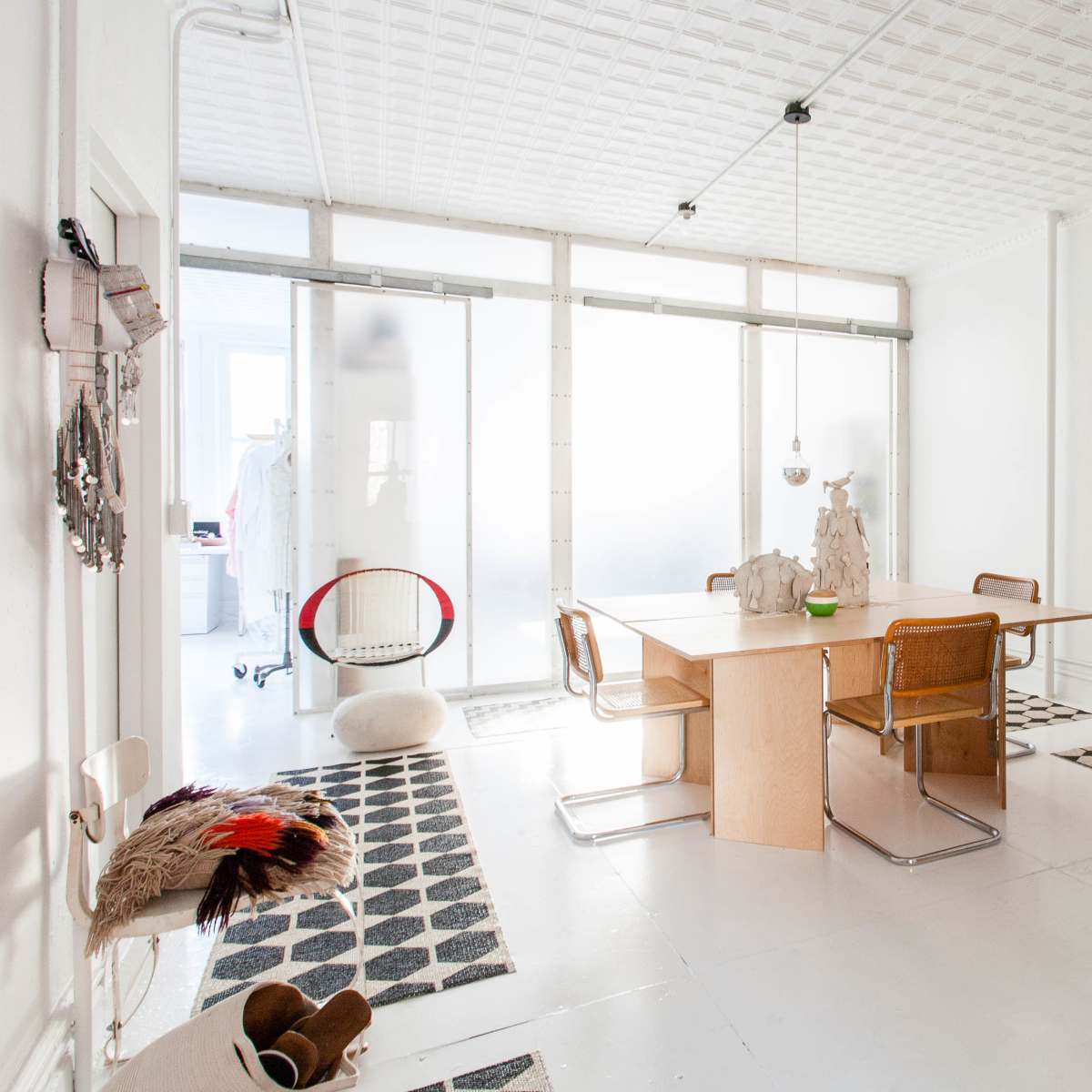A bright, minimalistic dining area featuring a large wooden table surrounded by chairs, with patterned rugs on the floor and a glass partition in the background.