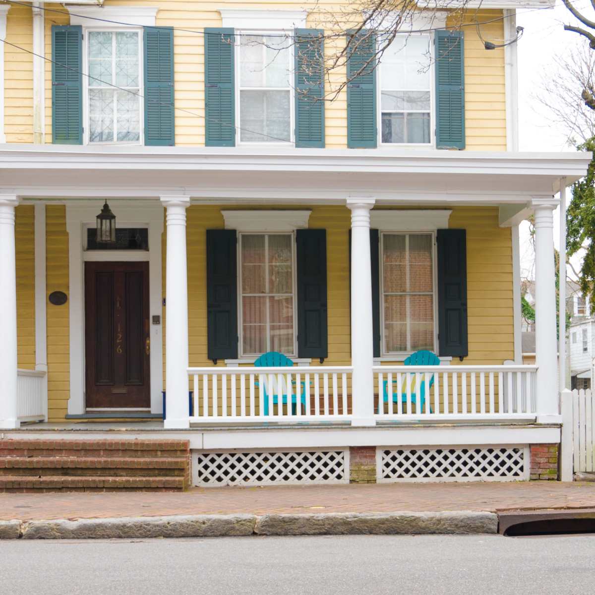 A two-story yellow house with green shutters, a front porch, and two blue chairs positioned on the porch.