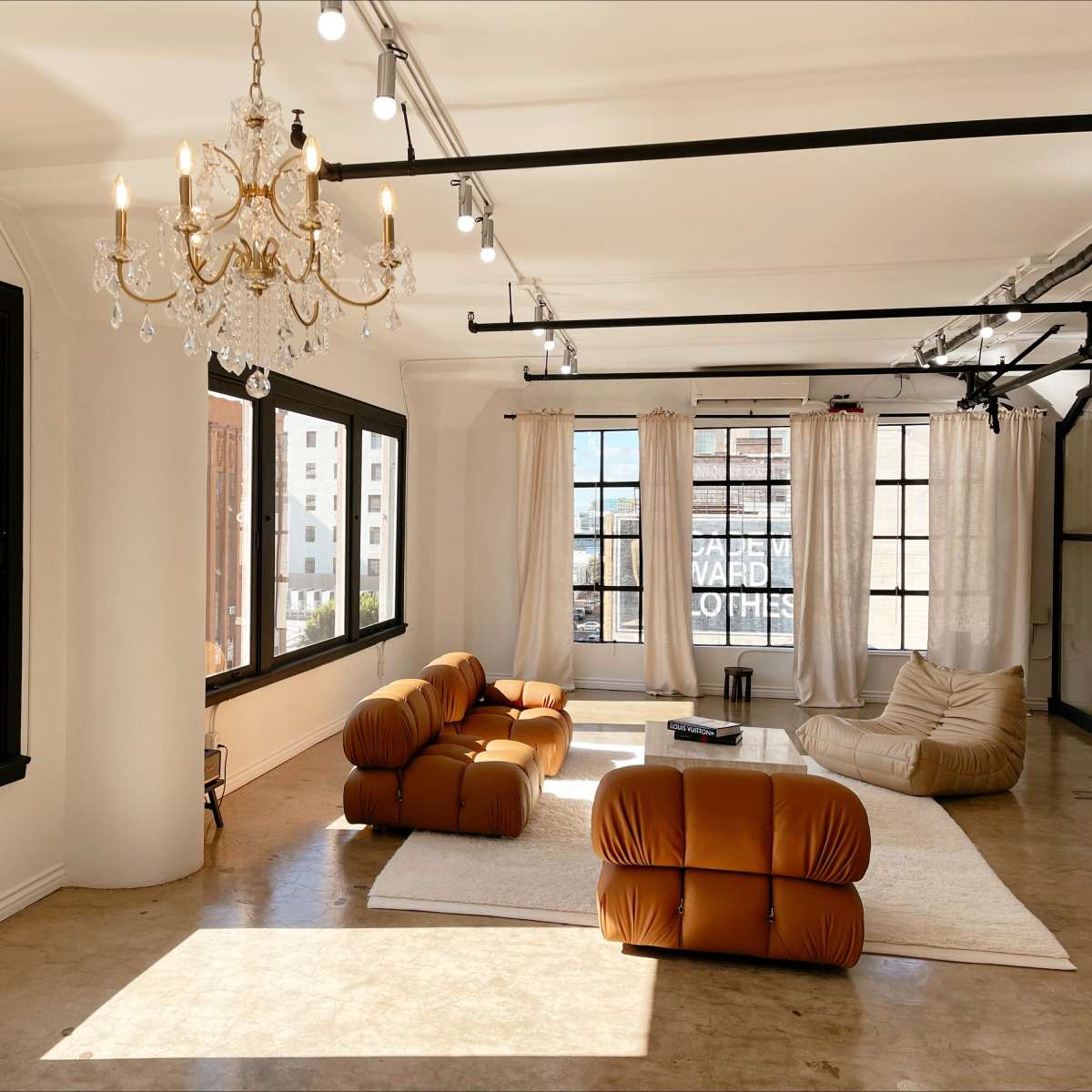 A modern, minimalist living space with large windows, a chandelier, and contemporary furniture arranged around a light-colored rug.