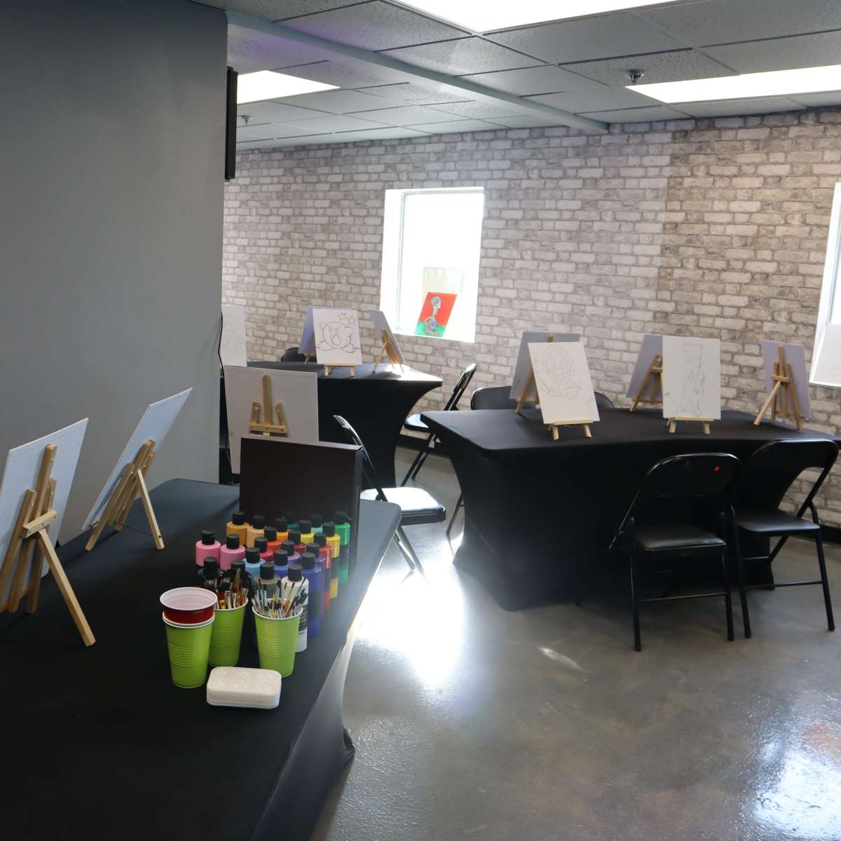 The scene shows a room set up for painting activities, featuring easels and tables covered with black cloth, along with supplies like cups and markers neatly arranged.