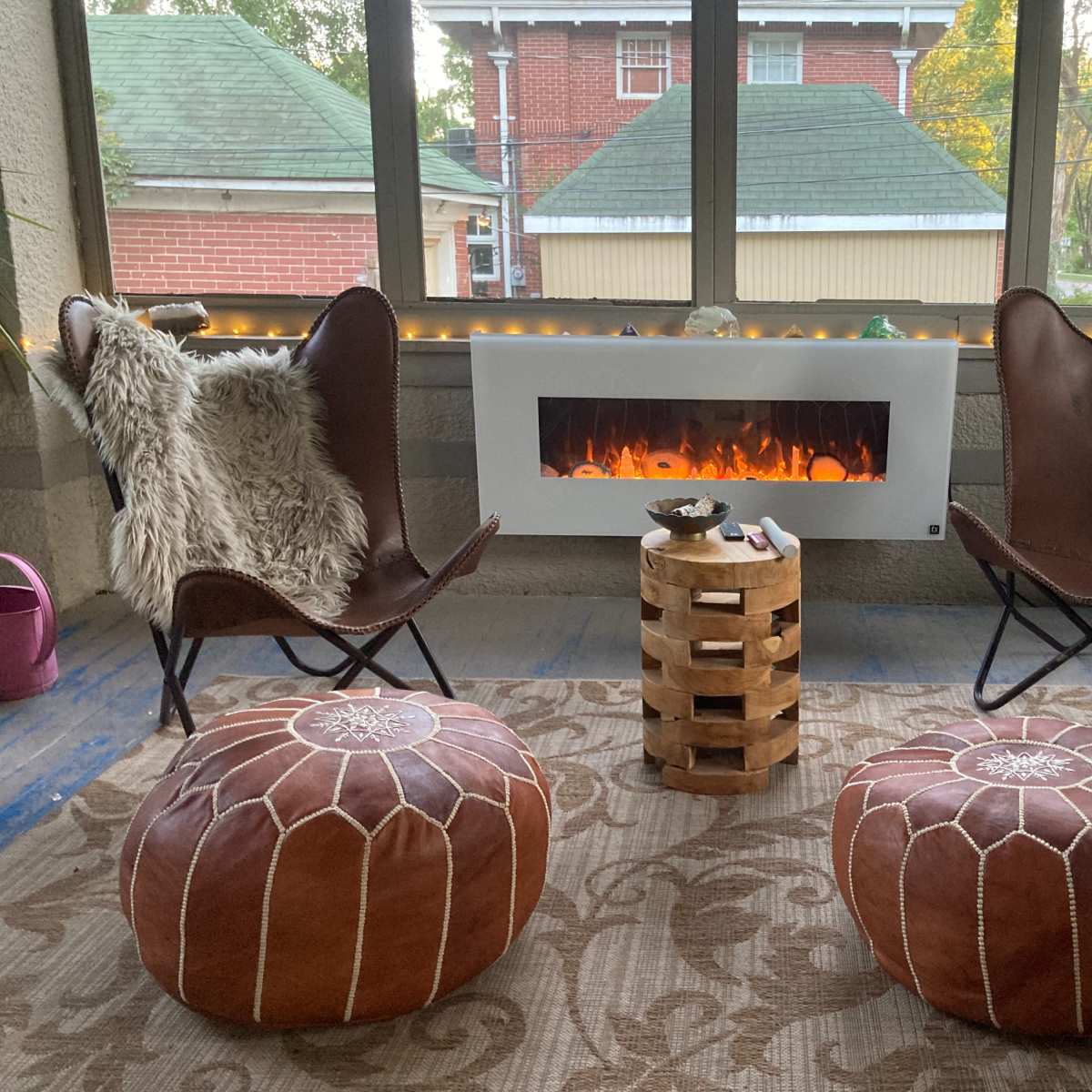 A cozy sunroom featuring two butterfly chairs, a wooden table, and a modern fireplace, with warm lighting in the background.