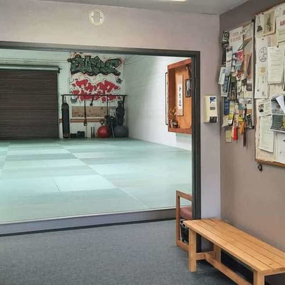 A hallway leading to a dojo, with a bulletin board on one wall and a bench in the foreground.