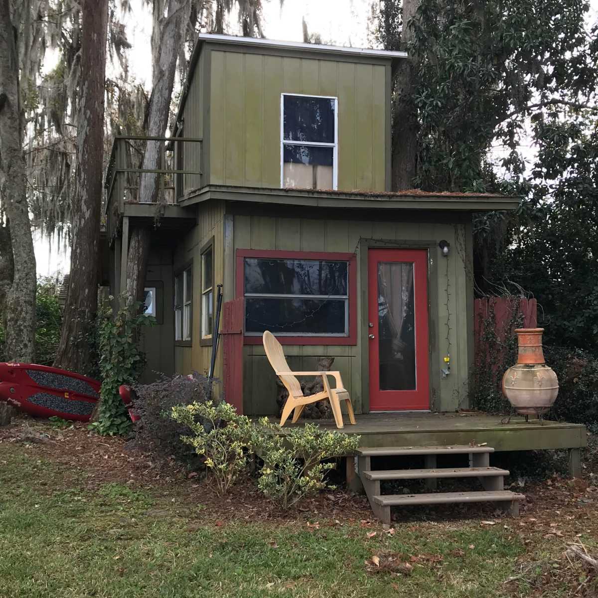 A small two-story green house with a red door and a wooden deck is surrounded by trees and grass.