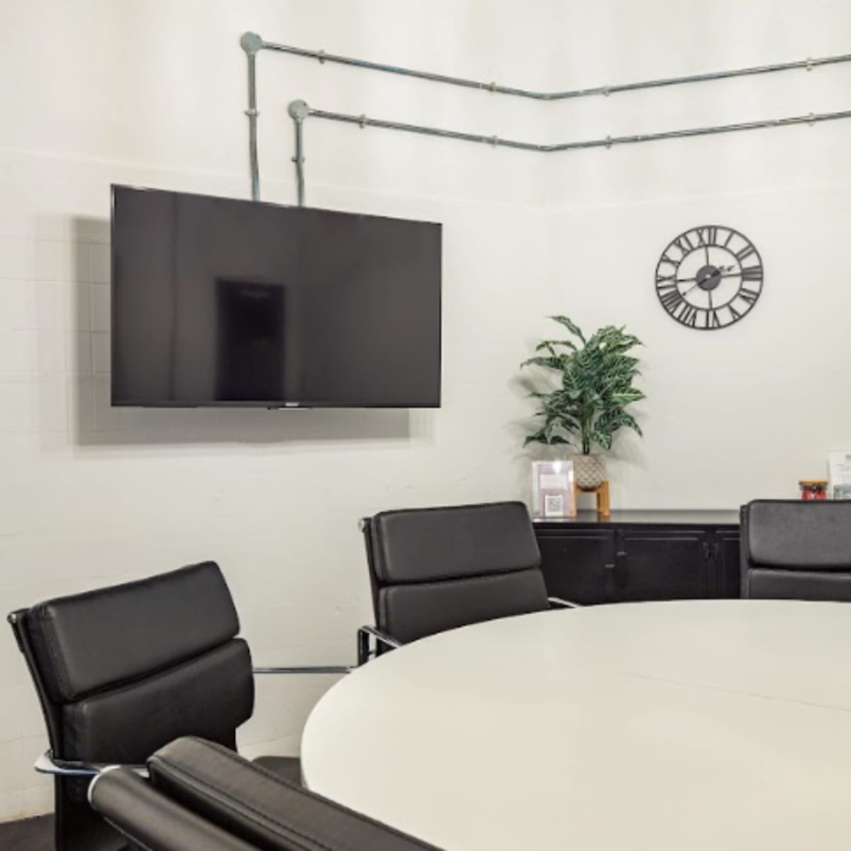 A modern conference room features a circular table, black leather chairs, a wall-mounted TV, and a clock, with industrial pipes and a potted plant.