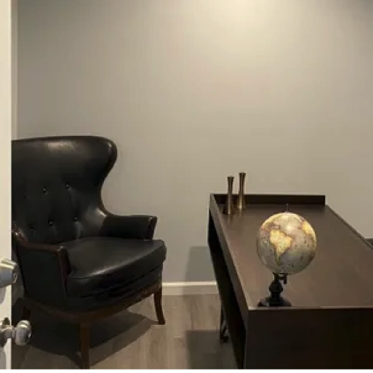 A small office space featuring a dark wooden desk, a globe on a stand, a black armchair, and a black office chair against a light gray wall.