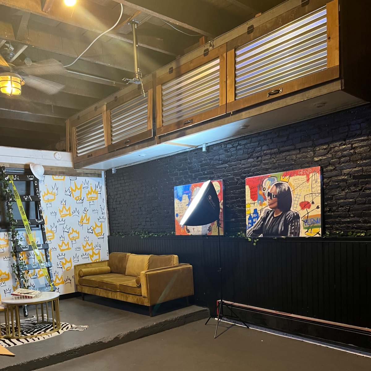 The room features a brown sofa, two framed artworks on a black wall, and decorative elements including a ladder and plants.