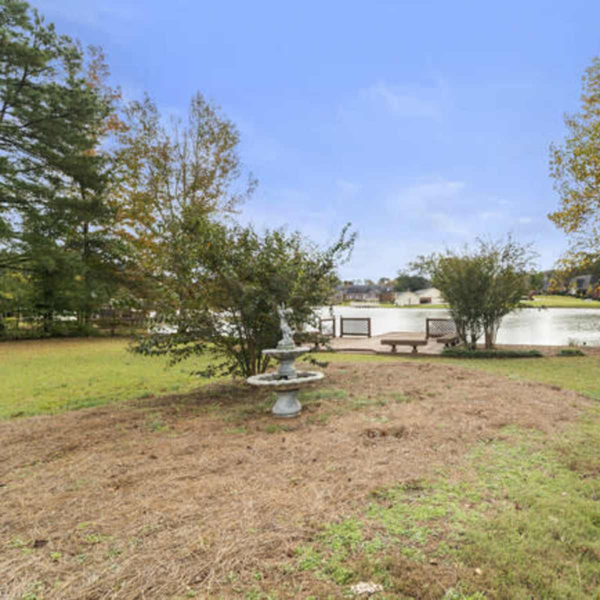 A grassy area with a birdbath and a wooden bench overlooks a calm lake surrounded by trees and houses.
