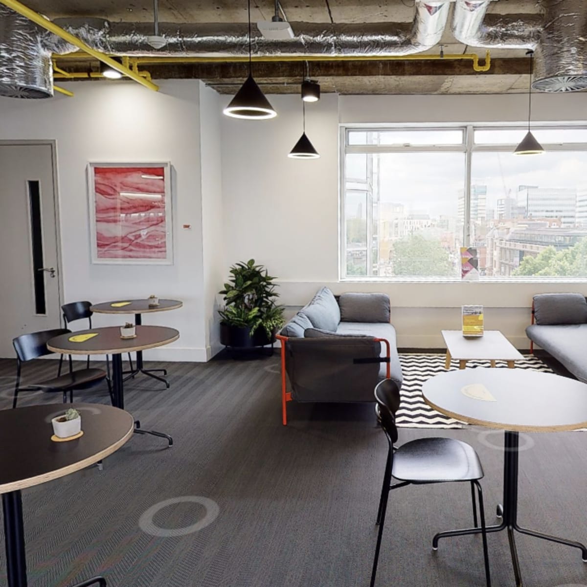 A modern office lounge with several round tables, a sofa, plants, and a large window overlooking the city.